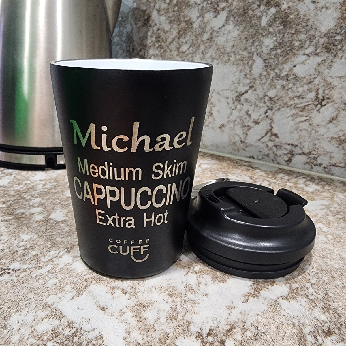 A black insulated coffee mug with a matching black lid sits on a speckled granite countertop. The mug is personalized with the name 'Michael' in green and cream text, along with 'Medium Skim CAPPUCCINO Extra Hot' and the brand 'COFFEE CUFF'.