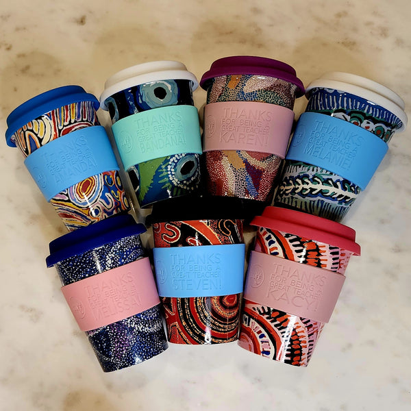 Six reusable travel mugs with colorful abstract and peacock-inspired patterns, arranged in a circle on a light beige marble surface. Each mug has a patterned body, a solid silicone sleeve in blue, pink, or black, and a patterned lid.