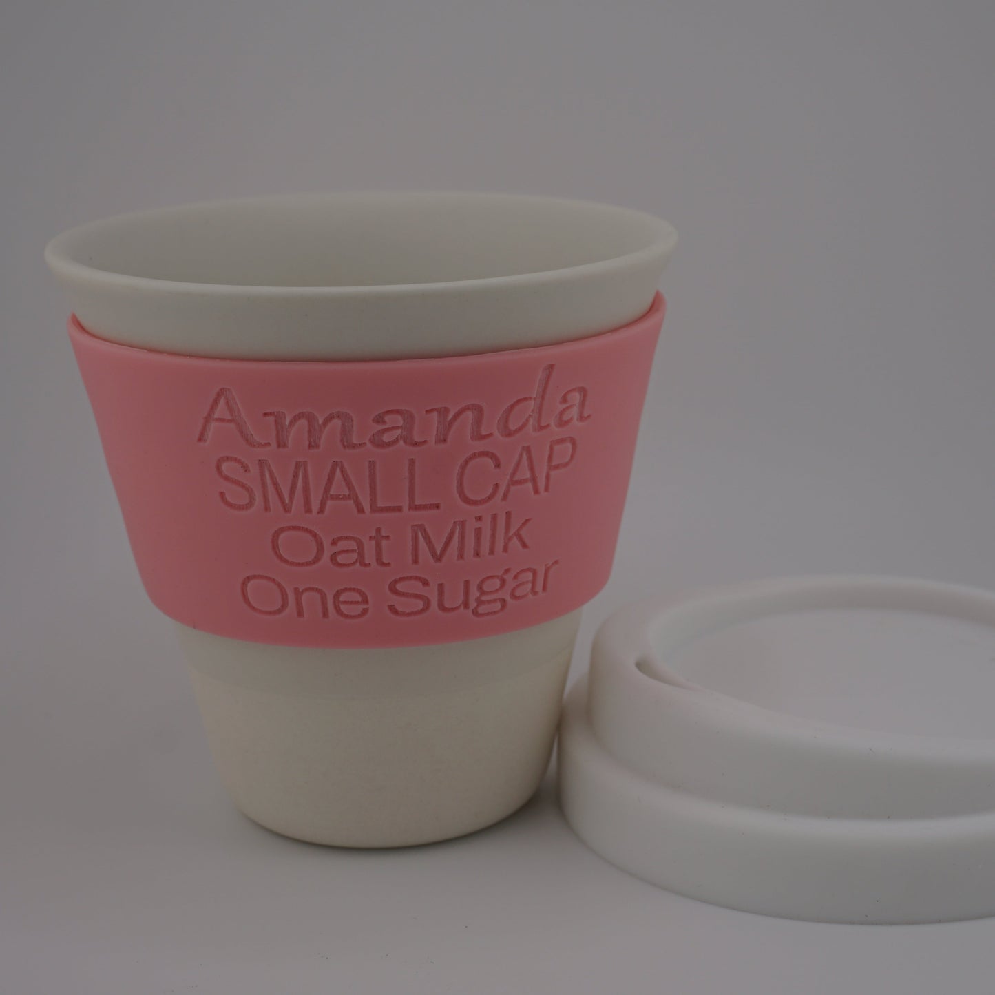 A reusable oat milk cup with a pink silicone sleeve printed with the text 'Amanda SMALL CAP Oat Milk One Sugar', shown next to its white lid on a light gray background.