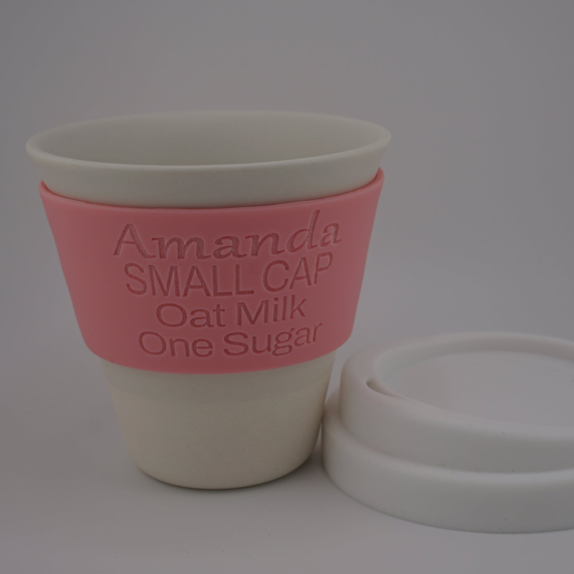 A reusable oat milk cup with a pink silicone sleeve printed with the text 'Amanda SMALL CAP Oat Milk One Sugar', shown next to its white lid on a light gray background.
