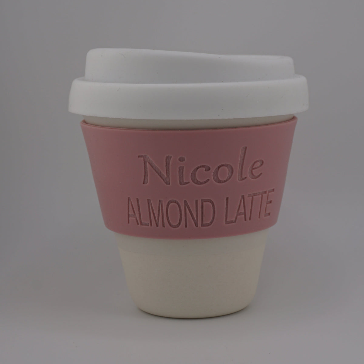 A reusable coffee cup with a white lid, a pink silicone sleeve embossed with the text 'Nicole ALMOND LATTE', and a cream-colored base, shown on a neutral gray background.