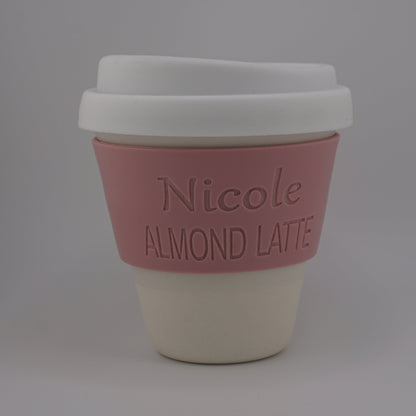 A reusable coffee cup with a white lid, a pink silicone sleeve embossed with the text 'Nicole ALMOND LATTE', and a cream-colored base, shown on a neutral gray background.
