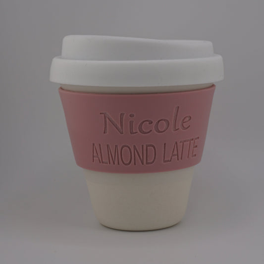 A reusable coffee cup with a white lid, a pink silicone sleeve embossed with the text 'Nicole ALMOND LATTE', and a cream-colored base, shown on a neutral gray background.