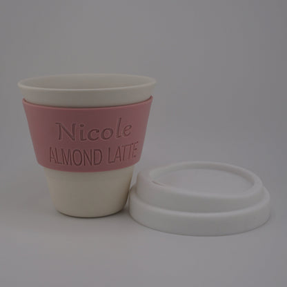 A reusable coffee cup with a pink sleeve printed with the text 'Nicole ALMOND LATTE', shown next to its white lid on a light gray background.