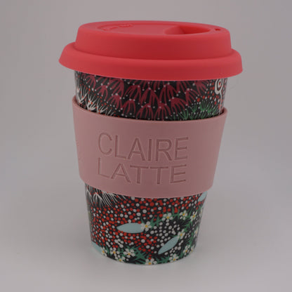 A reusable paper coffee cup with a pink silicone sleeve embossed with the text 'CLAIRE LATTE', a red lid, and a red, green, and white patterned lower section, set against a plain white background.