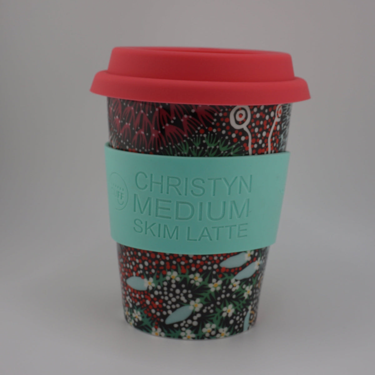 A reusable coffee cup with a red lid and a turquoise sleeve printed with the text 'CHRISTYN MEDIUM SKIM LATTE', set against a plain light gray background.