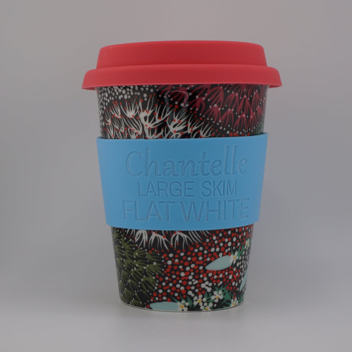 A reusable paper coffee cup with a red lid and a light blue sleeve printed with the text 'Chantelle LARGE SKIN HAND-WASHED'. The cup features a busy winter-themed pattern with red berries, green pine needles, and white snow.