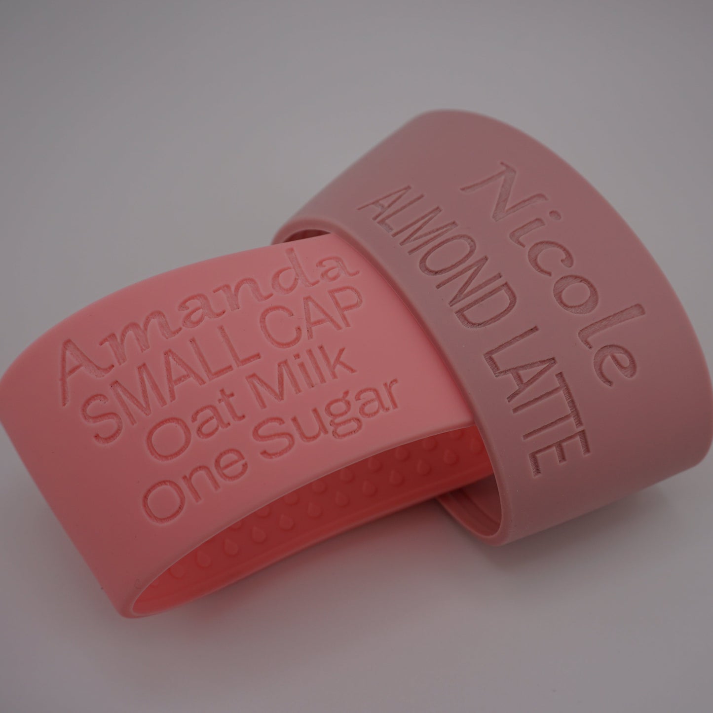 Two silicone coffee cup sleeves in shades of pink, one coral pink and one dusty rose pink, both embossed with text. One band reads 'Amanda SMALL CAP Oat Milk One Sugar' and the other reads 'Nicole ALMOND LATTE'.