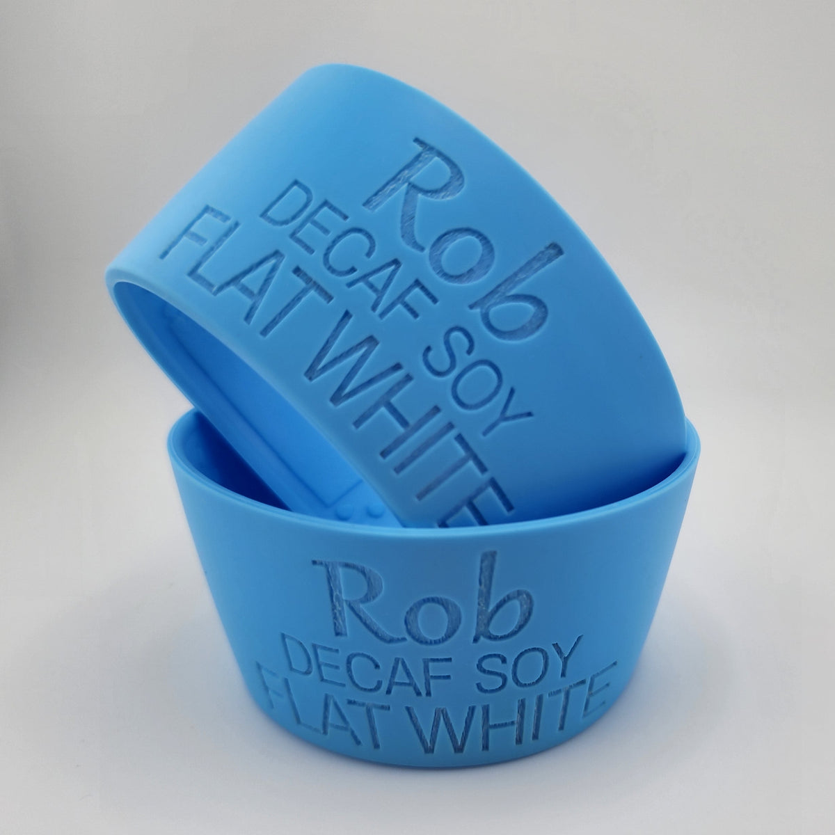 Two blue silicone coffee cups stacked on a white surface, embossed with the text 'Rob Decaf Soy Flat White'.