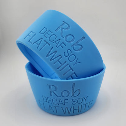 Two blue silicone coffee cups stacked on a white surface, embossed with the text 'Rob Decaf Soy Flat White'.