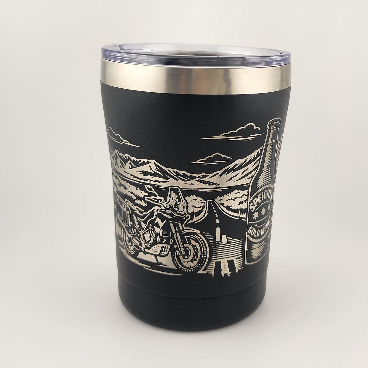 A black insulated tumbler with a clear lid and a silver stainless steel rim, featuring a white engraved graphic of a motorcycle and desert landscape. The tumbler is photographed on a plain white studio background.