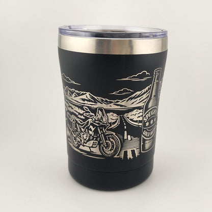 A black insulated tumbler with a clear lid and a silver stainless steel rim, featuring a white engraved graphic of a motorcycle and desert landscape. The tumbler is photographed on a plain white studio background.